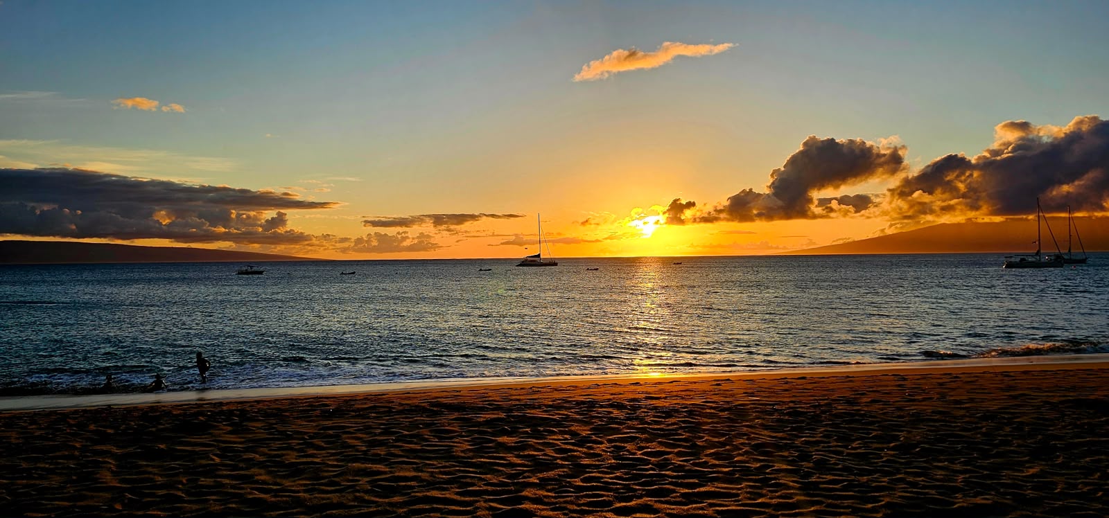 Sunset over the ocean with sailboats at anchor, people wading along the shoreline, and a sandy beach in the foreground